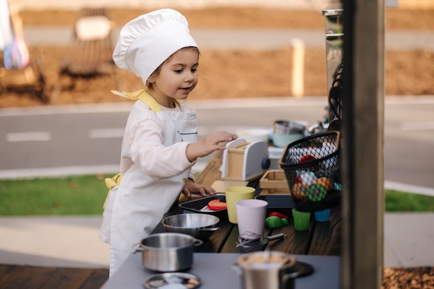 Adorabile bambina con cappotto e cappuccio da chef cuochi nella cucina giocattolo per bambini che giocano sui bambini piccoli