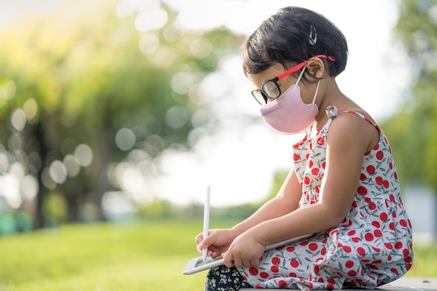 Adorabile bambina con maschera protettiva con gli occhiali seduta su una panchina del parco