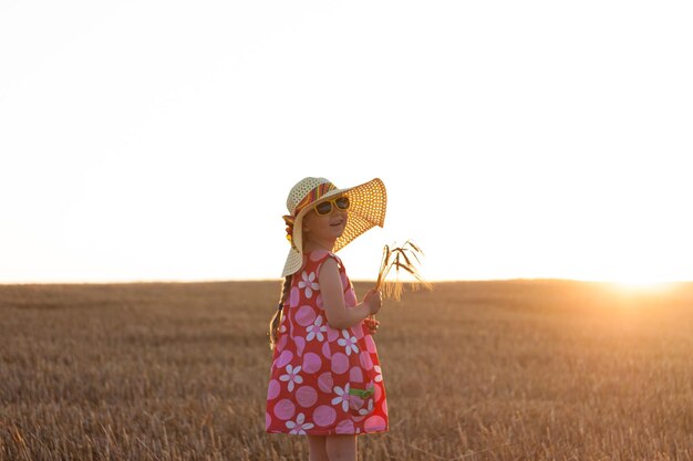 Adorabile bambina con un cappello di paglia e un abito estivo rosa nel campo di grano Bambino con lunghi capelli biondi sull'agricoltura al tramonto