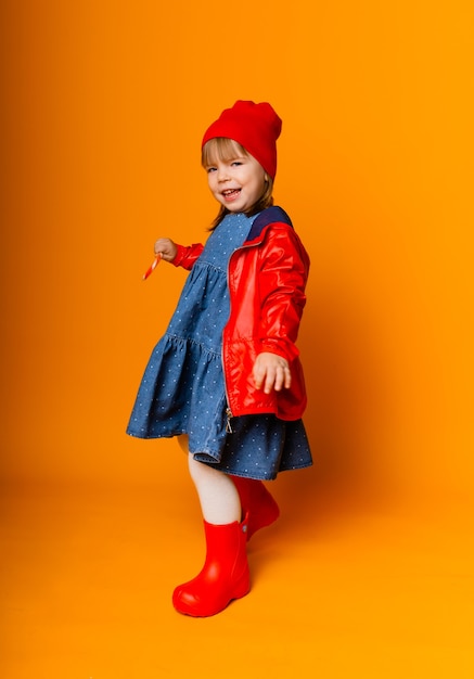 Adorabile bambina con una giacca rossa e stivali che tengono un lecca-lecca su uno sfondo giallo. concetto di autunno.
