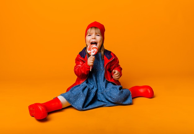 Adorabile bambina con una giacca rossa e stivali che tengono un lecca-lecca su uno sfondo giallo. concetto di autunno.