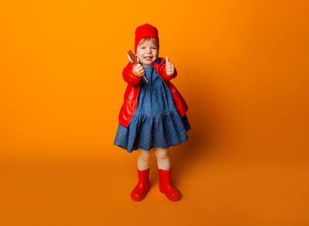 Adorabile bambina con una giacca rossa e stivali che tengono un lecca-lecca su uno sfondo giallo. concetto di autunno.