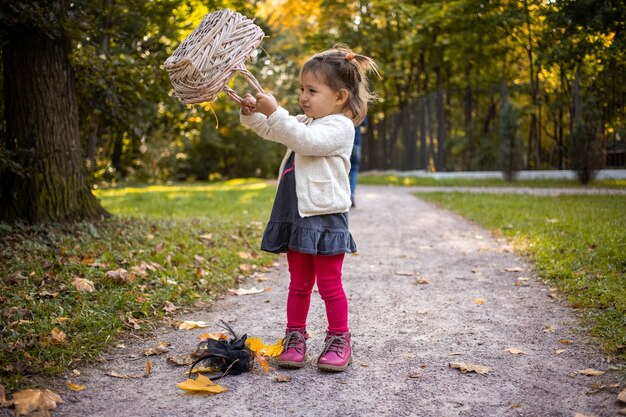 Adorabile bambina gioca con un cesto pieno di foglie nella foresta autunnale