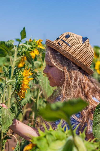 Adorabile bambina in abito estivo plaid cappello di paglia blu in un campo di campagna Bambino con i capelli lunghi odora di girasole