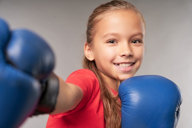 Adorabile bambina in guantoni da boxe che perfora l'aria e sorride mentre si trova in piedi su uno sfondo grigio