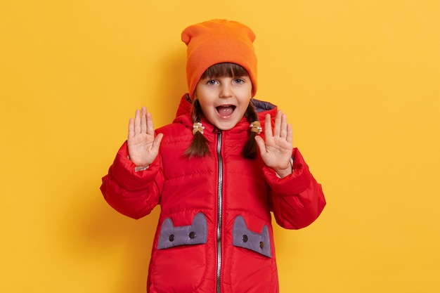 Adorabile bambina indossa giacca rossa casual e berretto arancione, tiene la bocca aperta, urlando qualcosa