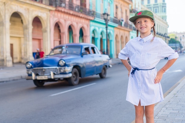 Adorabile bambina nella zona popolare di L'Avana Vecchia, Cuba. Ritratto di bambino, auto d'epoca classica americana