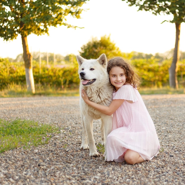 Adorabile bambina riccia con un grosso cane bianco nel parco Una bella bambina di 7 anni in abito rosa abbraccia il suo cane preferito durante una passeggiata estiva