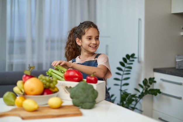 Adorabile bambina ridente e riccia si siede al tavolo in cucina, vicino alle verdure.