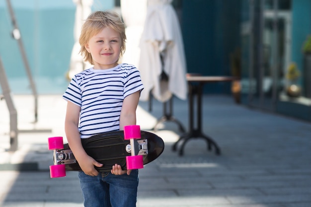 Adorabile bambino all'aperto. Bambino abbastanza allegro sveglio che tiene skateboard .. Ragazzo casuale in estate pattinaggio su uno skateboard.