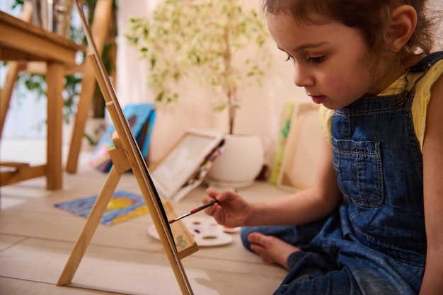 Adorabile bambino caucasico carino bambina seduta sul pavimento su un cavalletto in legno e disegno pittura su tela arte classe creatività bambini concetto di intrattenimento