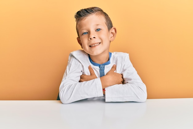 Adorabile bambino caucasico che indossa abiti casual seduto sul tavolo faccia felice sorridente con le braccia incrociate guardando la fotocamera persona positiva