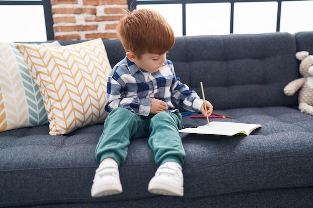 Adorabile bambino che disegna su un quaderno seduto sul divano a casa