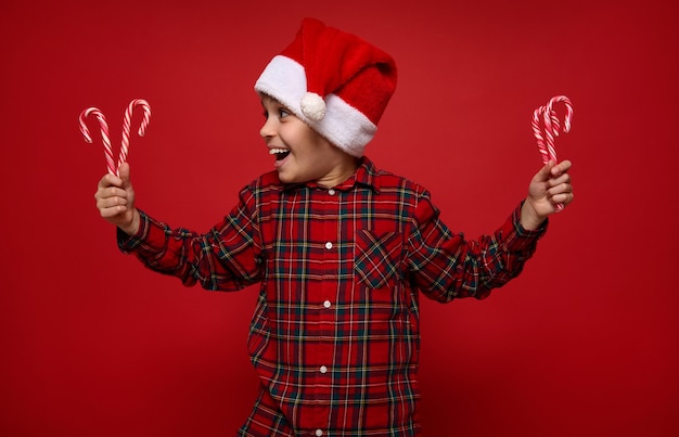 Adorabile bambino in camicia a quadri rossa e cappello di Babbo Natale guardando lecca-lecca di Natale, bastoncini di zucchero a strisce nelle sue mani, isolato su sfondo di colore rosso con spazio per la copia per la pubblicità