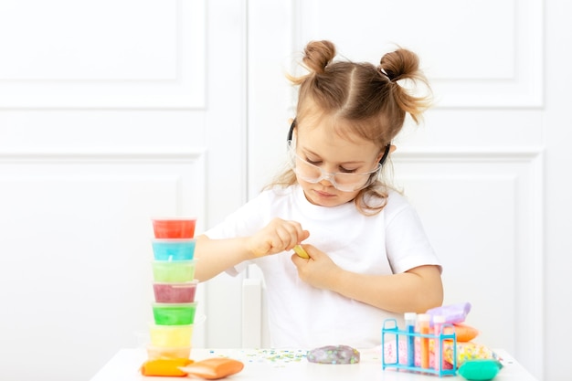 Adorabile bambino in una maglietta bianca con due code si siede al tavolo con occhiali protettivi e facendo esperimenti, producendo una melma soffice da ingredienti multicolori
