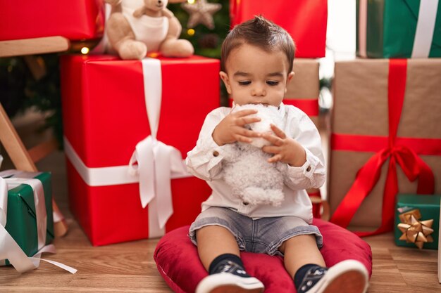 Adorabile bambino ispanico che abbraccia l'orsacchiotto seduto sul pavimento vicino ai regali di Natale a casa