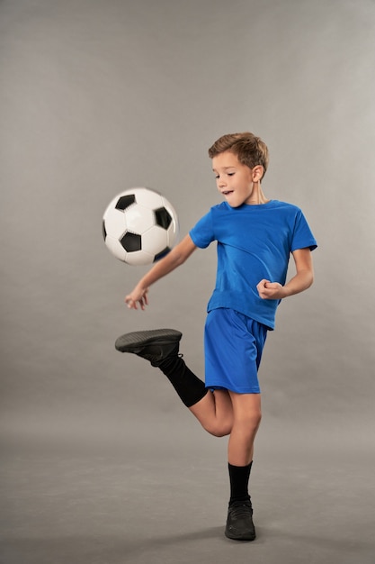 Adorabile bambino maschio in pantaloncini che fa acrobazie di calcio su sfondo grigio