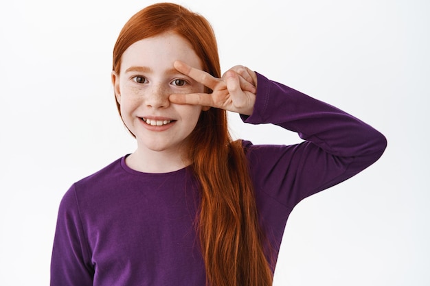 Adorabile bambino, ragazza allo zenzero con lunghi capelli naturali e lentiggini, sorridente ampiamente e mostrando il segno di pace v sull'occhio, bambino felice che sembra ottimista, sfondo bianco