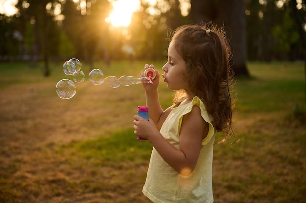 Adorabile bella bambina, che soffia bolle di sapone al tramonto, godendosi piacevoli momenti all'aperto nel prato. I raggi del sole cadono attraverso sfere di bolle trasparenti con riflessi iridescenti.