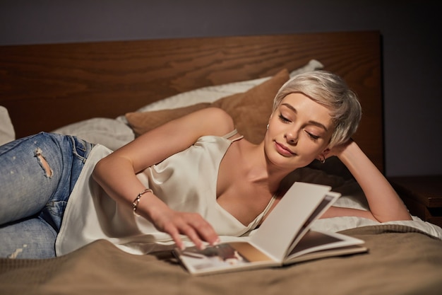 Adorabile donna caucasica con i capelli corti biondi sdraiata sul letto guardando le foto sulla rivista, godendosi il tempo di relax di notte, riposati