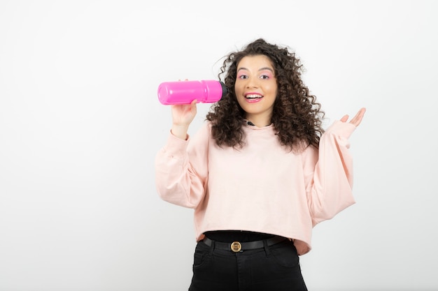 Adorabile donna riccia con bottiglia d'acqua rosa in piedi sopra il bianco.