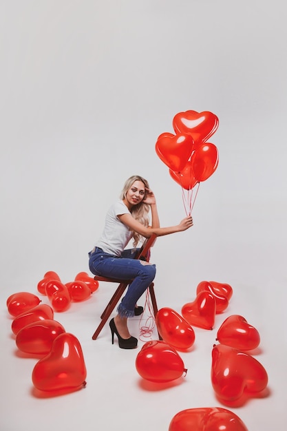 Adorabile giovane ragazza bionda in jeans e t-shirt bianca in posa con palloncini a forma di cuore su sfondo bianco il giorno di San Valentino concetto