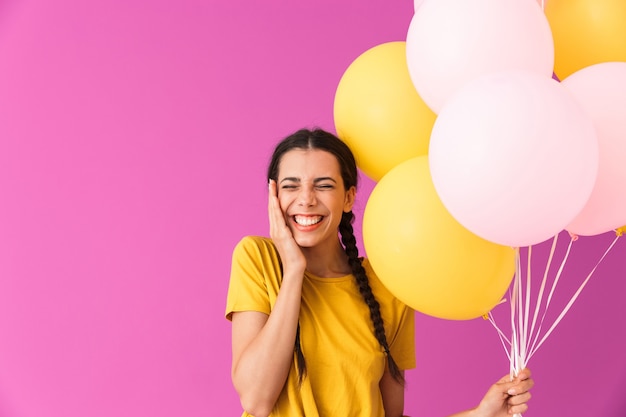 Adorabile giovane ragazza bruna che tiene in mano un mazzo di mongolfiere mentre sta in piedi isolata sul rosa, celebrando