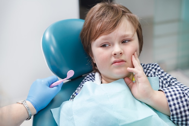 Adorabile giovane ragazzo con mal di denti, seduto in poltrona odontoiatrica