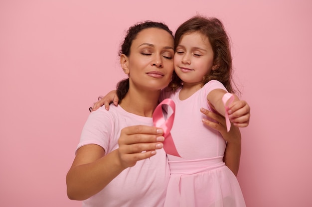 Adorabile madre donna di razza mista abbraccia una bambina in abiti rosa e mostra un nastro di raso rosa alla fotocamera, simbolo della Giornata del cancro al seno nel mese di ottobre. Sostegno ai malati di cancro e ai sopravvissuti