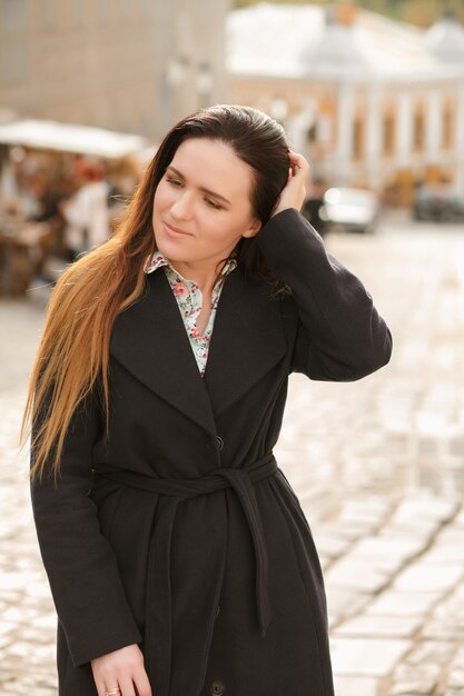 Adorabile modella bruna con i capelli lunghi che indossa un cappotto blu alla moda e una camicia di cotone in posa alla giornata di sole