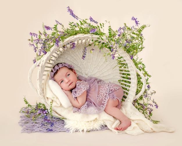 Adorabile neonata che indossa un bel vestito e una corona sdraiata nel cesto con decorazioni vegetali che si tengono per mano sotto le guance in studio. Bambino neonato carino con fiori