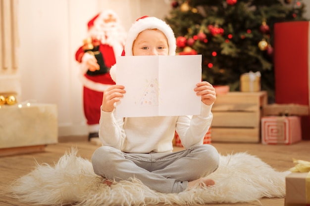 Adorabile piccolo artista che indossa un cappello di Babbo Natale seduto sul pavimento e si nasconde dietro il suo nuovo capolavoro con sopra un albero di Natale.