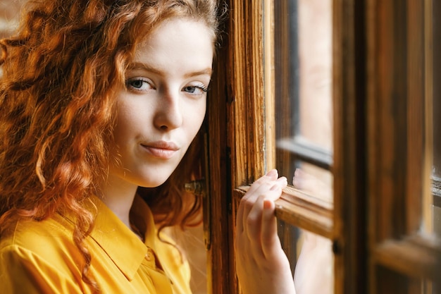 Adorabile ragazza adolescente riccia zenzero con la camicia gialla in piedi vicino alla finestra e in posa alla telecamera all'interno del loft