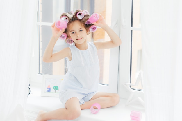 Adorabile ragazza carina con bigodini sui capelli, si siede sul davanzale della finestra a casa, andando ad avere un'acconciatura riccia