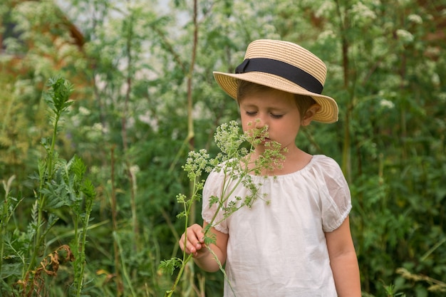 Adorabile ragazza carina in un abito boho bianco con un cappello annusando fiori di campo nel villaggio