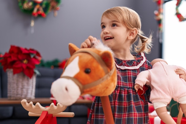 Adorabile ragazza caucasica che tiene baby doll e cavallo giocattolo in piedi accanto alla decorazione natalizia a casa
