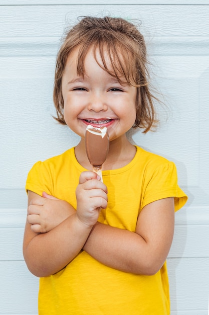 Adorabile ragazza dai capelli biondi che mangia il gelato, indossa una camicia gialla appoggiata su sfondo bianco