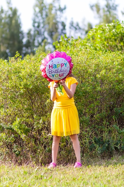 Adorabile ragazza in abito giallo con palloncino colorato festivo di buon compleanno a forma di fiore. Bambino felice all'aperto