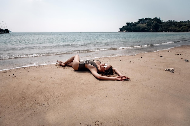 Adorabile ragazza in forma con petto espressivo in bikini con scollo aperto è sdraiata su una spiaggia sabbiosa vicino al mare, grandangolo