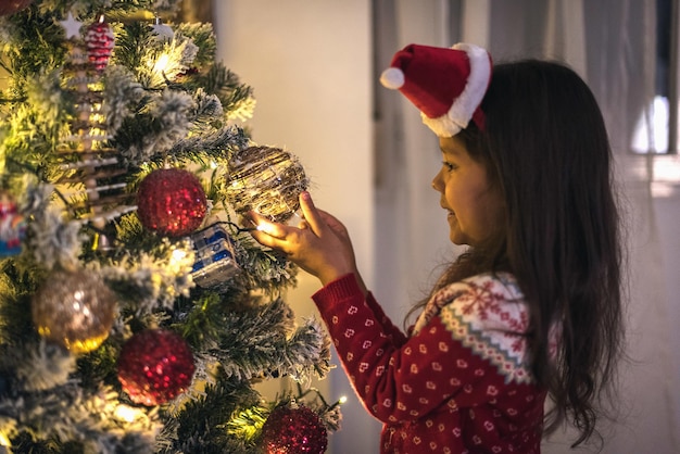 Adorabile ragazza ispanica in un maglione natalizio e un cerchio per la testa in posa vicino all'albero di Natale decorato