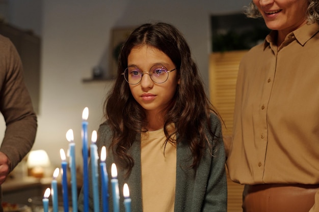 Adorabile ragazza preadolescente con gli occhiali che guarda le candele della menorah in fiamme