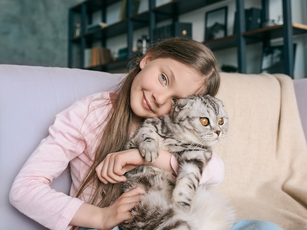 Adorabile ragazza sorridente carina che abbraccia il gatto grigio scozzese sul divano di casa Animali domestici amicizia a casa