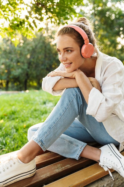 Adorabile ragazza vestita casualmente seduta su una panchina nel parco cittadino, ascoltando musica con cuffie wireless