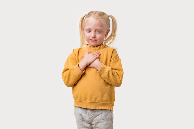 Adorabile ragazzina caucasica con i capelli biondi in una felpa gialla in piedi con le mani piegate su uno sfondo bianco