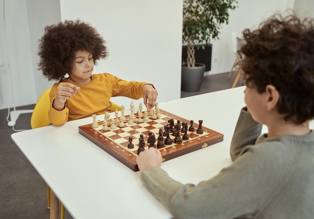 Adorabile ragazzino con i capelli afro che fa una mossa mentre gioca a scacchi con il suo amico seduto