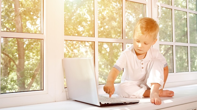 Adorabile ragazzino seduto sul davanzale di una grande finestra bianca e premendo il pulsante sul laptop, luce solare