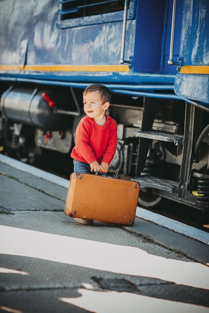 Adorabile ragazzino vestito con un maglione rosso su una stazione ferroviaria vicino al treno con una vecchia valigia marrone retrò. Pronto per le vacanze. Giovane viaggiatore sulla piattaforma.