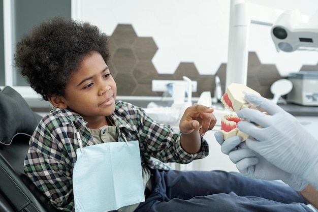 Adorabile ragazzo africano che indica i denti falsi tenuti dalle mani guantate dello stomatologo