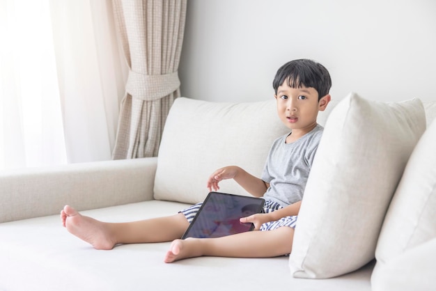 Adorabile ragazzo asiatico felice che indossa una camicia grigia e pantaloncini a righe bluewhite si diverte a giocare con il suo tablet su un divano color crema guardando lo schermo del cellulare