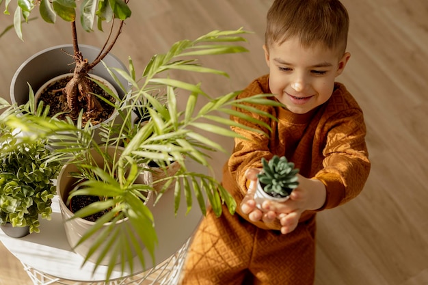 Adorabile ragazzo carino che si prende cura delle piante d'appartamento a casa Un piccolo aiuto nell'attività per il tempo libero domestico Concetto di giardinaggio domestico Camera accogliente Colori della terra Abbigliamento casual